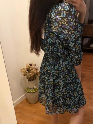 Blommig kortklänning med puffärm - Superfin kortklänning med svart botten och blommigt mönster i blått, lila och grönt. Klänningen har puffiga långärmar och markerad midja med resår. Tunt, lätt material som ger en luftig känsla. Perfekt för dig som gillar romantiska vibes och färg.