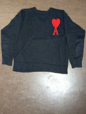 Svart stickad tröja från Ami - Svart stickad tröja från Ami Alexandre Mattiussi med röd hjärtlogga och bokstaven A på bröstet. Tröjan har rund hals, ribbstickade muddar och avslut. Perfekt för dig som gillar stilrena plagg med en twist.