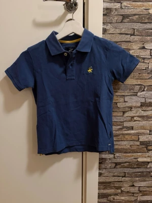 Marinblå pikétröja med broderad logga - Snygg marinblå pikétröja från U.S. Polo Assn. med klassisk krage, korta ärmar och broderad gul logga på bröstet. Tröjan är i mjuk bomull och har knappar vid halsen. Perfekt för en avslappnad och stilren look.