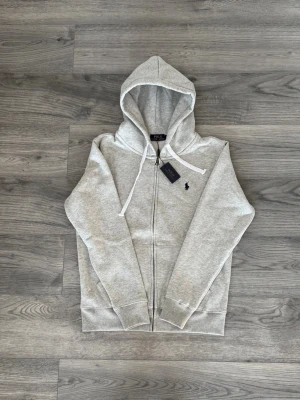 Grå zip-hoodie från Polo Ralph Lauren - Snygg ljusgrå hoodie med dragkedja från Polo Ralph Lauren. Klassisk design med huva och vita snören, ribbade muddar och fickor framtill. Liten broderad logga på bröstet. Perfekt för en chill och stilren look.