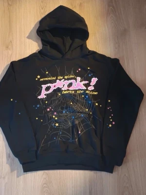 Spider hoddie  - Cool svart hoodie med huva och tryck framtill. Motivet har ett stort spindelnät, färgglada stjärnor och texten 'nevermind the spider, PINK! heres the slime' i rosa, vitt och gult. Perfekt för dig som gillar unika och lekfulla plagg.
