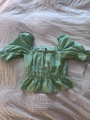 Grön rutig off-shoulder topp - Söt grön och vit rutig topp med off-shoulder design och puffärmar. Smockad midja och volang nertill ger en feminin siluett. Perfekt för sommardagar och picknickar. Materialet känns lätt och luftigt.💕💕storlek kan variera mellan 32-34 alltså xxs 
