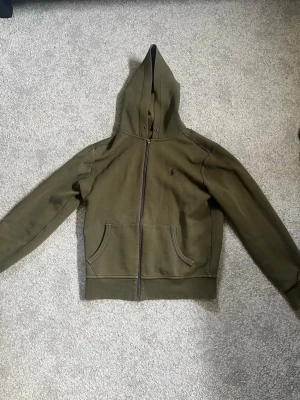 Olivgrön hoodie från Polo Ralph Lauren - Snygg olivgrön hoodie från Polo Ralph Lauren med dragkedja framtill och två stora fickor. Klassisk huva med snörning och diskret logga på bröstet. Tillverkad i mjuk bomullsblandning som är skön att ha på sig.