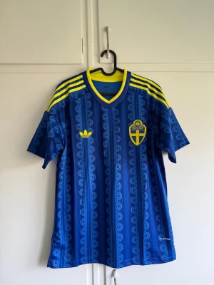 Sverige fotbollströja borta 2026  - Säljer en blå fotbollströja från Adidas med Sveriges landslagsemblem på bröstet. Tröjan har gula detaljer, tre gula ränder på axlarna och ett mönster i blå nyanser. Tillverkad i lätt och ventilerande material, perfekt för träning eller match.