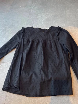 Svart blus med volangdetaljer - Svart blus med diskret prickigt mönster och volangdetaljer vid axlarna. Blusen har rund halsringning och långa ärmar. Perfekt att styla med jeans eller kjol för en trendig look.