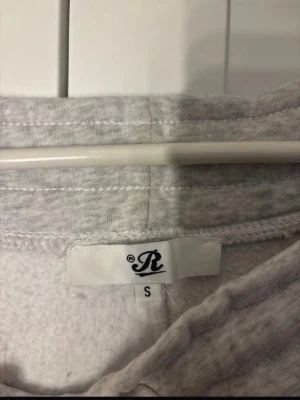 Ljusgrå mjukisbyxor med dragsko - Ljusgrå mjukisbyxor från R med dragsko i midjan och fickor på sidorna. Byxorna har en klassisk rak passform och är tillverkade i mjukt, fleeceliknande material för extra komfort. Perfekta för en avslappnad stil. Dåligt tvättade men det löser sig med en tvätt