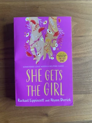 She Gets the Girl pocketbok - En färgstark pocketbok med lila omslag och illustrationer av två tjejer bland blommor. Boken är skriven av Rachael Lippincott och Alyson Derrick och handlar om kärlek, vänskap och att våga ta chanser. Perfekt för dig som gillar romantik och ungdomslitteratur.