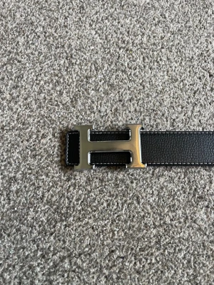 Svart skärp med H-spänne - Snyggt svart skärp med H-format metallspännet. Skärpet är gjort i slät skinn med vita sömmar längs kanterna för en clean och exklusiv look. Perfekt accessoar för att lyfta vilken outfit som helst. Skärpet är 110 cm långt och priset går att diskutera