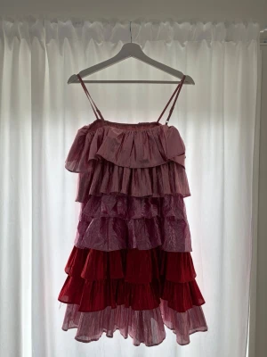 Rosa och röd volangklänning Collective - Säljer en superfin kortklänning från Collective The Label med flera lager volanger i rosa och rött. Klänningen har smala axelband och ett glittrigt material som ger en cool look. Perfekt för dig som vill sticka ut och älskar färg!