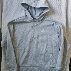 Grå hoodie från Adidas - Säljer en stilren grå hoodie från Adidas med vit logga på bröstet. Hoodien har huva med dragsko och är tillverkad i mjukt bomullsmaterial. Perfekt för dig som gillar sportig och avslappnad stil.