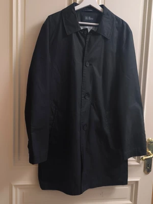 Svart trenchcoat från Oscar Jacobson - Stilren svart trenchcoat från Oscar Jacobson med klassisk krage och knäppning framtill. Jackan har raka ärmar och diskreta sidofickor. Materialet är slätt och ger ett elegant intryck. Perfekt för dig som vill ha en tidlös och snygg look.