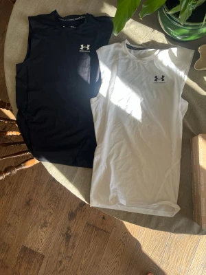 Under Armour träningslinnen SVART+VIT - Två ärmlösa träningslinnen från Under Armour i svart och vitt. Slimmad passform med rund hals och diskret logga på bröstet. Tillverkade i ett lätt och stretchigt funktionsmaterial som andas, perfekta för gym eller löpning.