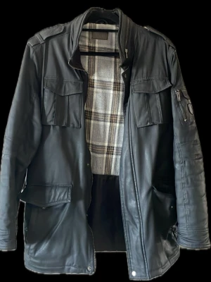 Skinnjacka Modell M65/militär  - Snygg svart skinnjacka från JF med flera stora fickor framtill, axelklaffar och dragkedja. Insidan har rutigt foder i grått och beige. Detaljer som patch på ärmen och dragkedjor ger jackan en cool look.