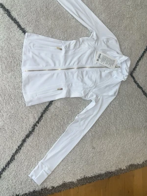 Lululemon  - Snygg vit lululemon med hel dragkedja framtill och två fickor med dragkedja. Koftan har lång ärm och figurnära passform, perfekt för träning eller utomhusaktiviteter. Tillverkad i ett stretchigt och lätt material.