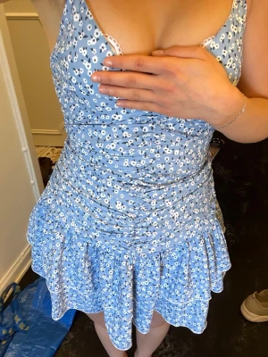 Blå blommig kort klänning med volang - Söt blå kort klänning med vita små blommor som tyvärr blivit för lite. V-ringning och smala axelband som är justerbara. Dragkedja i ryggen. Klänningen har volang nertill och lätt rynkade detaljer över magen. Perfekt till sommaren. Klänningen är i mycket bra skick, som ny!