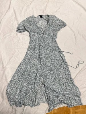 Blommig omlottklänning från Lindex - Supersöt lång omlottklänning från Lindex i vitt och grönt med blommigt mönster. Klänningen har korta ärmar, volangkant nertill och knytband i midjan. Perfekt för vår och sommar när du vill känna dig extra fin.