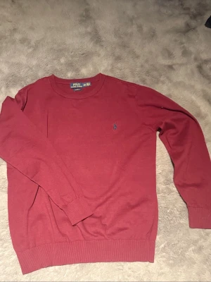 Röd stickad tröja från Polo Ralph Lauren - Snygg röd stickad tröja från Polo Ralph Lauren med klassisk blå broderad logga på bröstet. Tröjan har rund halsringning, långa ärmar och ribbade muddar vid ärmslut och nederkant. Perfekt för lager på lager och enkel att matcha.