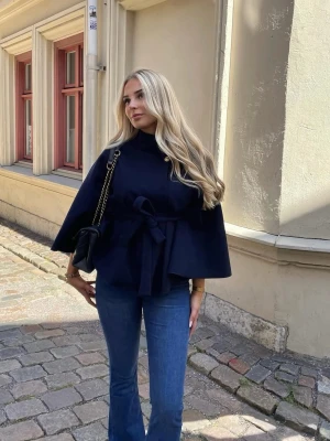 Mörkblå cape-kappa med knytskärp - Snygg mörkblå cape-kappa med hög krage och knytskärp i midjan. Kappan har vida ärmar och en elegant, stilren look som passar perfekt till jeans eller klänning. Materialet ser ut att vara ullblandning och modellen är trendig och modern.