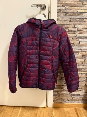 Lila och röd pufferjacka Quechua - Snygg pufferjacka från Quechua i lila och rött med svarta dragkedjor och huva. Jackan har två fickor med dragkedja och quiltad design. Perfekt för kalla dagar och riktigt skön att ha på sig. Materialet är lätt och isolerande.