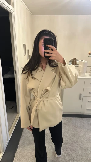 Beige kappa med knytskärp - Stilren beige kappa med knytskärp i midjan och breda slag. Rak passform och långa ärmar ger en avslappnad men chic look. Perfekt för dig som gillar minimalistisk stil. Materialet ser ut att vara mjukt och följsamt, troligen ullblandning.