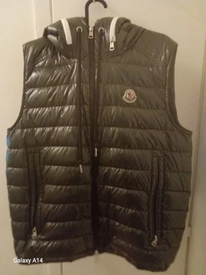 Svart dunväst från Moncler med luva  - Snygg svart dunväst från Moncler med glansig finish och avtagbar luva. Västen har två dragkedjefickor, luva och Moncler-logga på bröstet. 