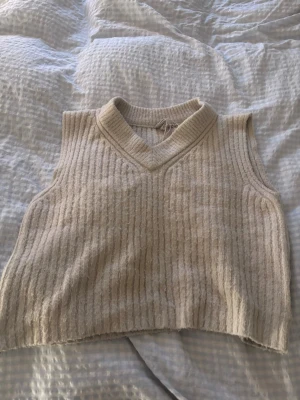 Beige ribbad stickad väst - Säljer en beige stickad väst med ribbad struktur och v-ringad hals. Västen är croppad och har en mjuk, mysig känsla som passar perfekt för lager på lager. Enkel och stilren design utan knappar eller dragkedja.💕