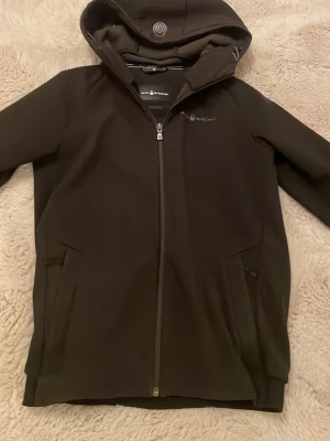 Svart vindjacka från Sailracing - Svart vindjacka från Sail racing med hel dragkedja, två sidofickor och justerbar huva. Jackan har en stilren design och är tillverkad i ett mjukt, vindtätt material som passar perfekt för blåsiga dagar. Snygg logga på bröstet och detaljer vid huvan.