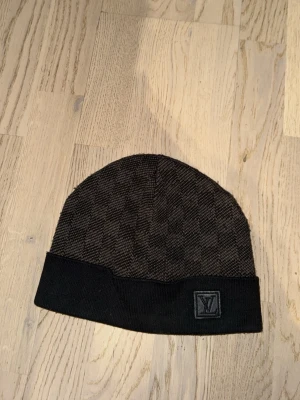 mössa - Snygg svart mössa med diskret rutmönster och uppvikt kant. Märkets logotyp sitter framtill på kanten. Perfekt accessoar för att hålla värmen med stil.