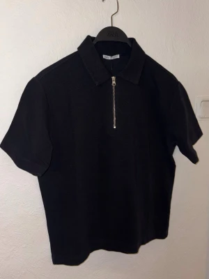 Svart polotopp med dragkedja från Zara - Svart polotopp från Zara med kort ärm och klassisk krage. Toppen har en snygg silverfärgad dragkedja framtill och är tillverkad i ett mjukt, stickat material. Perfekt att styla med jeans eller kjol för en trendig look.