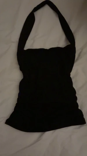 Svart halterneck linnetopp - Trendig svart linnetopp med halterneck och breda axelband som kan korsas eller knytas i nacken. Toppen är ärmlös och har en tight passform som framhäver siluetten. Perfekt för att skapa en snygg och stilren look.