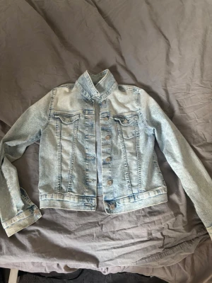 Ljusblå jeansjacka från H&M - Snygg ljusblå jeansjacka från H&M med klassisk knappstängning framtill och två bröstfickor med lock. Jackan har en rak passform och coola sömdetaljer som ger en trendig look. Perfekt att slänga över en hoodie eller t-shirt.