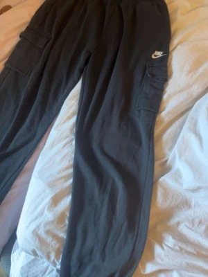 Svarta cargobyxor från Nike - Säljer ett par svarta cargobyxor från Nike med klassisk logga på låret. Byxorna har flera stora fickor på sidorna och resår i midjan. Perfekta för en avslappnad streetstil och tillverkade i mjukt material.
