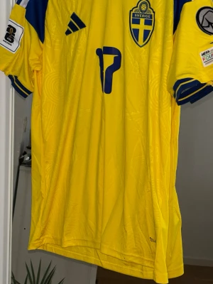 Sverige VM fotbollströja Gyökeres 17 - Gul och blå officiell Sverige fotbollströja från Adidas med tryck Gyökeres 17 på ryggen. Tröjan har broderad landslagssköld, Adidas-logga och detaljerade mönster i tyget. Korta ärmar med blå ränder och UEFA-patch på ärmen. Perfekt för match eller supporterhäng. Tröjan fick man som present men den är lite för stor så tänker sälja