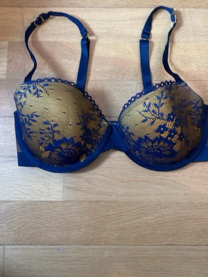 Stella McCartney BH mycket bra - Snygg bh i mörkblått med guldfärgad botten och blå spets med blommönster. Justerbara axelband och bygel för extra stöd. Kuporna är vadderade och har fina spetsdetaljer. Perfekt för dig som gillar en elegant och feminin stil.