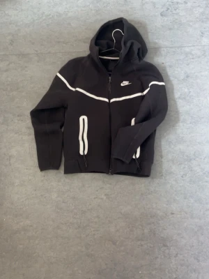 Svart Nike hoodie med vit detalj - Svart hoodie från Nike med dragkedja och vita detaljer längs bröstet och vid fickorna. Klassisk huva och två fickor framtill. Perfekt för en sportig och avslappnad stil. Nike-logga broderad på bröstet.