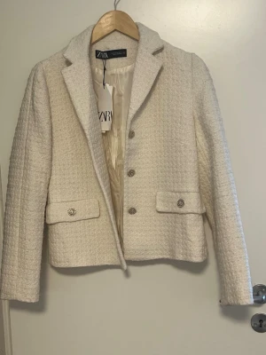 Krämvit kavaj från Zara - Stilren krämvit kavaj från Zara i strukturerat tyg med klassisk krage och dekorativa silverknappar. Kavajen har två fickor framtill med lock och är helfodrad för en lyxig känsla. Perfekt för att lyfta din outfit med en elegant touch.