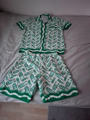 Grönt och vitt set med mönster - Säljer ett matchande set från Cider i storlek M. Setet består av en kortärmad skjorta och shorts i vitt och grönt med grafiskt mönster och gröna kanter. Materialet är polyester och känns lätt och svalt, perfekt för sommaren.