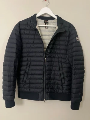 Svart dunväst från Colmar Originals - Snygg svart dunväst från Colmar Originals med quiltad design och ribbade muddar. Västen har dragkedja framtill, två sidofickor med dragkedja och en ljusgrå insida. Perfekt för lager-på-lager och håller dig varm under kyliga dagar.