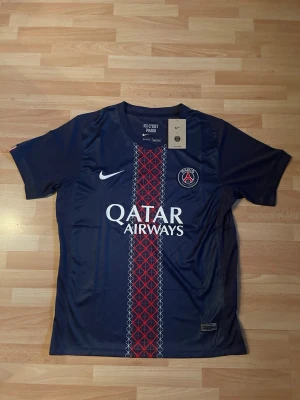 PSG matchtröja Nike mörkblå - Mörkblå Paris Saint-Germain matchtröja från Nike med klubbmärke på bröstet och Qatar Airways-tryck. Tröjan har korta ärmar och ett rött och vitt mönster längs mitten. Tillverkad i lätt och ventilerande material, perfekt för fotbollsträning.