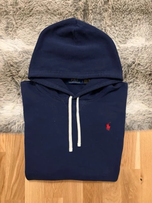 Mörkblå hoodie från Polo Ralph Lauren - Snygg mörkblå hoodie med vit dragsko och klassisk röd Polo-logga broderad på bröstet. Hoodien har en stor huva och är gjord i mjukt bomullsmaterial som känns riktigt skönt. Perfekt för dig som vill ha en stilren och tidlös look.