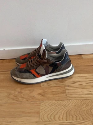 Kamouflage sneakers med orange detaljer - Sneakers med grymt kamouflagemönster i grönt, brunt och orange. Ovandel i mocka och textil, bruna skosnören och vit sula med svart undersida. Snygga detaljer och cool streetstil, perfekt för dig som vill sticka ut.