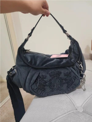 Juicy Couture hobo axelväska - Mörkblå handväska från Juicy Couture i mjuk velour med broderad logga framtill. Väskan har silverfärgade metalldetaljer, dragkedja upptill och ett dekorativt hjärthänge. Både axelrem och handtag finns för olika stilar.