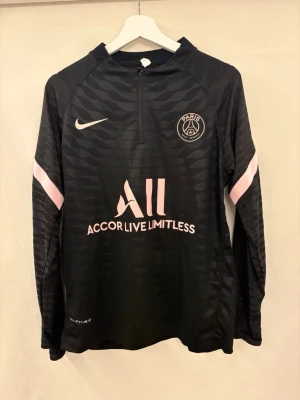 Svart PSG träningströja Nike - Svart långärmad träningströja från Nike för Paris Saint-Germain med rosa detaljer och klubbmärke på bröstet. Halvlång dragkedja framtill, tryckta loggor och texten 'Accor Live Limitless' samt 'Visit Rwanda' på ryggen. Tillverkad i funktionsmaterial. Helt oanvänd på grund av fel storlek.