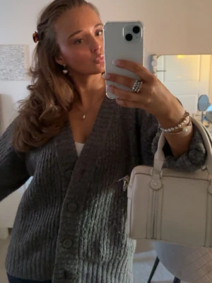 Grå stickad cardigan med stora knappar - Mysig grå stickad cardigan med djup v-ringning och stora knappar framtill. Modellen är lite oversized med långa ärmar och ribbstickade detaljer. Perfekt att slänga över en topp för en chill och trendig look.