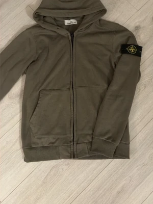Olivgrön hoodie från Stone Island - Snygg olivgrön hoodie från Stone Island med dragkedja framtill och klassisk logotyp-patch på vänster ärm. Hoodien har två stora fickor fram och ribbade muddar vid ärmslut och nederkant. Perfekt för en avslappnad streetwear-look.