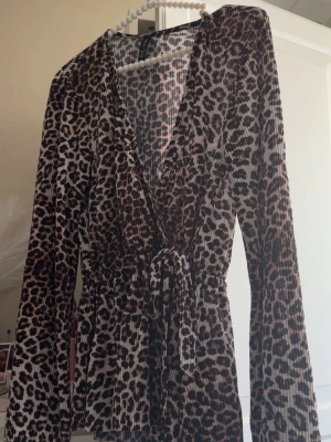 Leopardmönstrad omlottopp med knyt - Säljer en leopardmönstrad omlottopp med knyt i midjan. Toppen har lång ärm, v-ringning och är i ett stretchigt, mjukt material som känns bekvämt. Perfekt för dig som vill sticka ut med ett djurmönster och gillar trendiga detaljer.