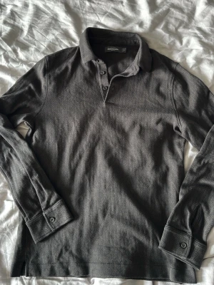 Grå henley-skjorta från Dressmann - Snygg grå långärmad henley-skjorta från Dressmann med diskret fiskbensmönster. Skjortan har knappar vid halsen och muddar med knappar vid ärmsluten. Perfekt för en stilren och avslappnad look.