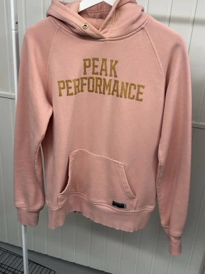 Rosa hoodie från Peak Performance -  Rosa hoodie från Peak Performance med stor guld logga framtill. Lite gulnad i kragen!