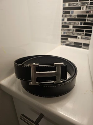 Svart skärp med H-spänne - Snyggt svart bälte med vit kontrastsöm och ett stort, silverfärgat H-format metallspänne. Perfekt accessoar för att lyfta din outfit med en stilren och modern touch. DM vid intresse!