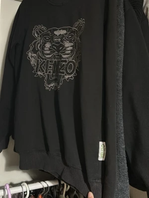 Svart sweatshirt med tiger från Kenzo - Svart sweatshirt från Kenzo med ikoniskt tigerbroderi och logga på bröstet. Tröjan har rund halsringning, ribbade muddar och en liten Kenzo-tag på sidan. Tillverkad i mjukt bomullsmaterial och har en avslappnad passform.